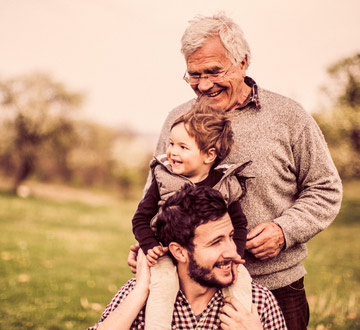 Ein älterer Mann steht hinter einem jüngeren Mann, der ein Kind auf den Schultern trägt. Alle lächeln und befinden sich im Freien.