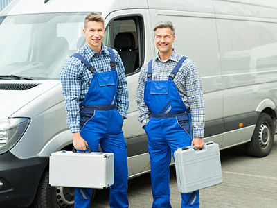 Zwei Handwerker in blauen Overalls stehen neben einem Lieferwagen und halten Werkzeugkisten.